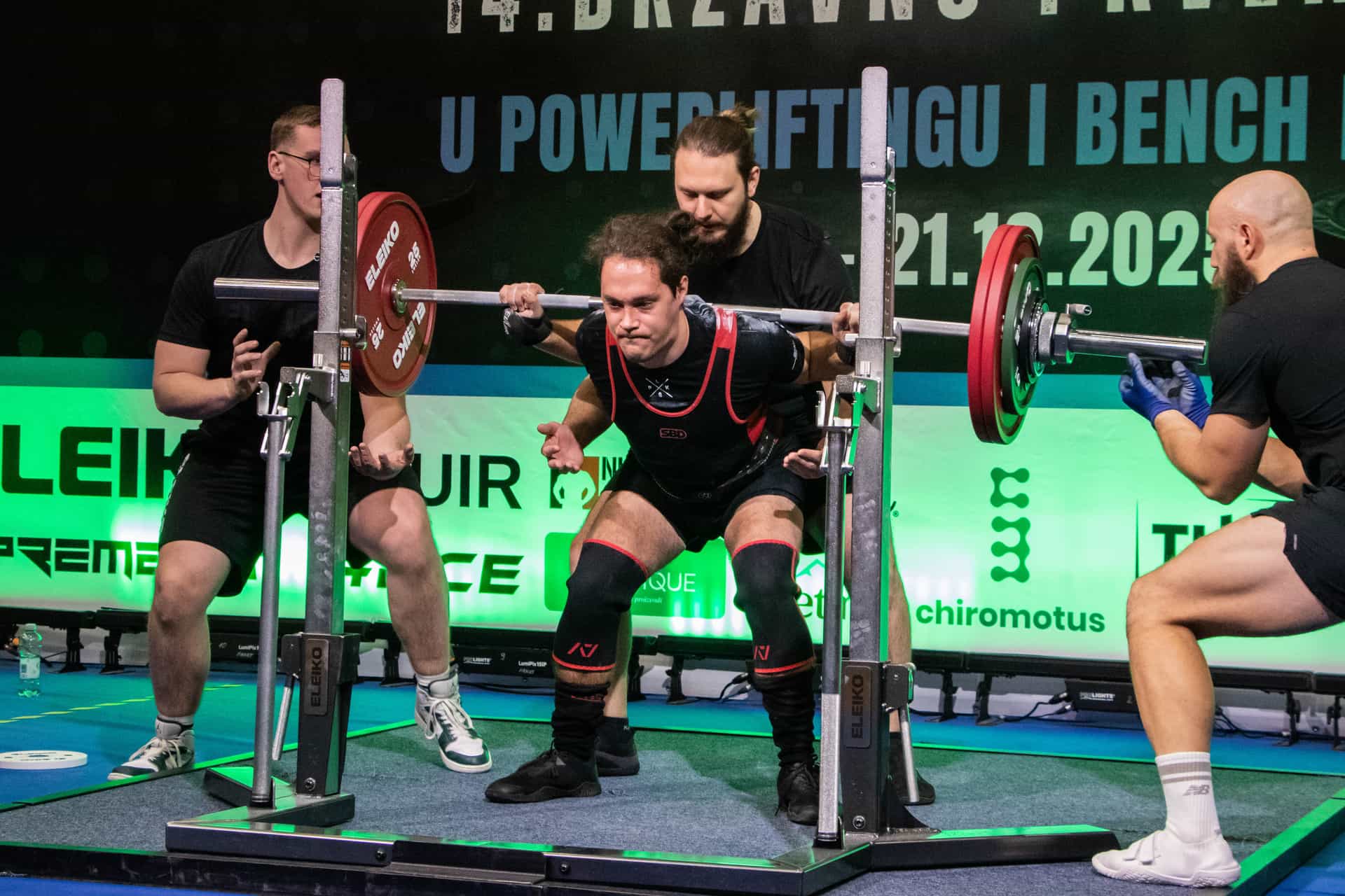 Natjecatelj izvodi čučanj na državnom prvenstvu u powerliftingu, uz pomoćnike na platformi