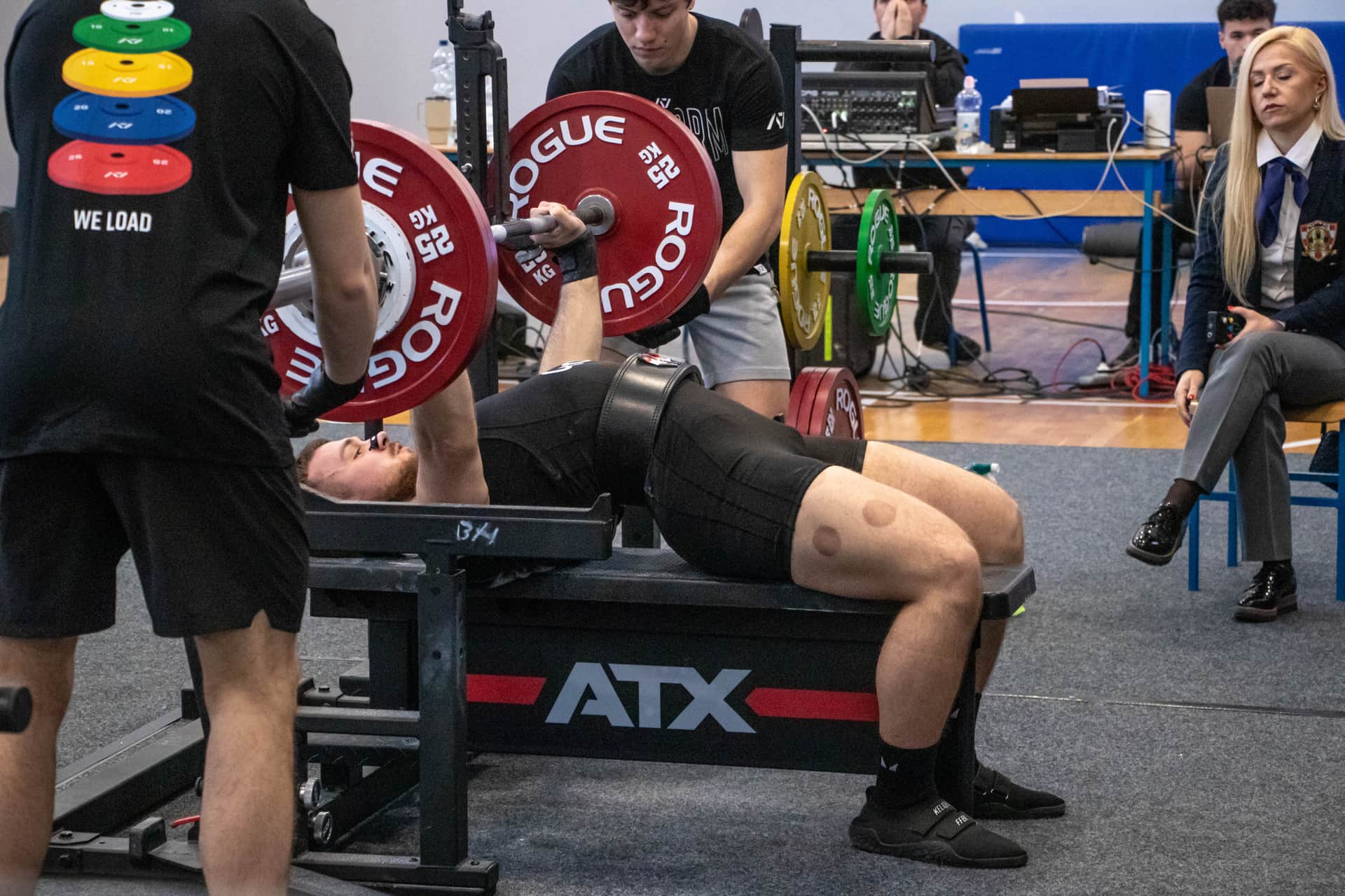 Natjecatelj izvodi bench press na natjecateljskoj klupi, uz pomoćnike na platformi