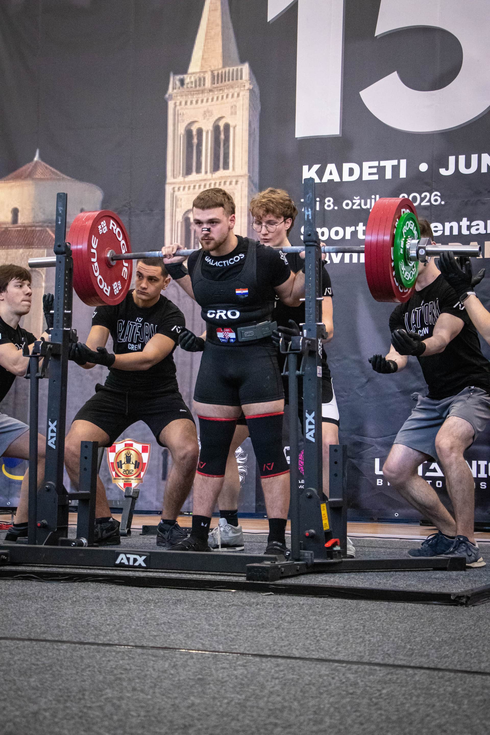 Natjecatelj sa šipkom na leđima izvodi čučanj na powerlifting natjecanju, uz pomoćnike sa strane